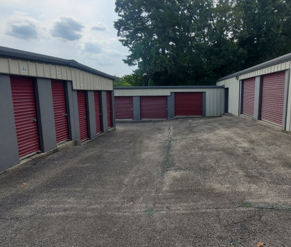 Storage units with red doors, a gravel lot, and trees under a cloudy sky.