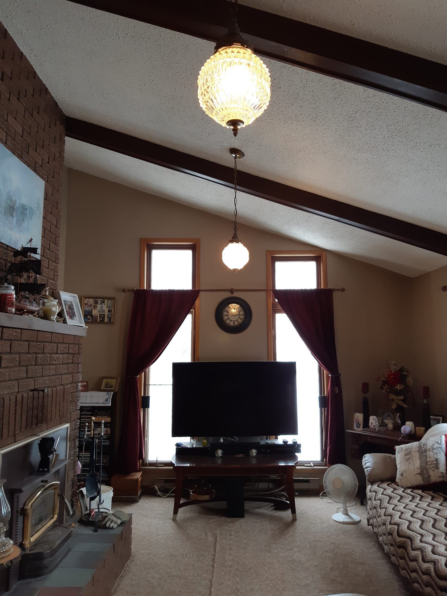 Living room with brick fireplace, large TV, windows, and reddish-brown curtains.