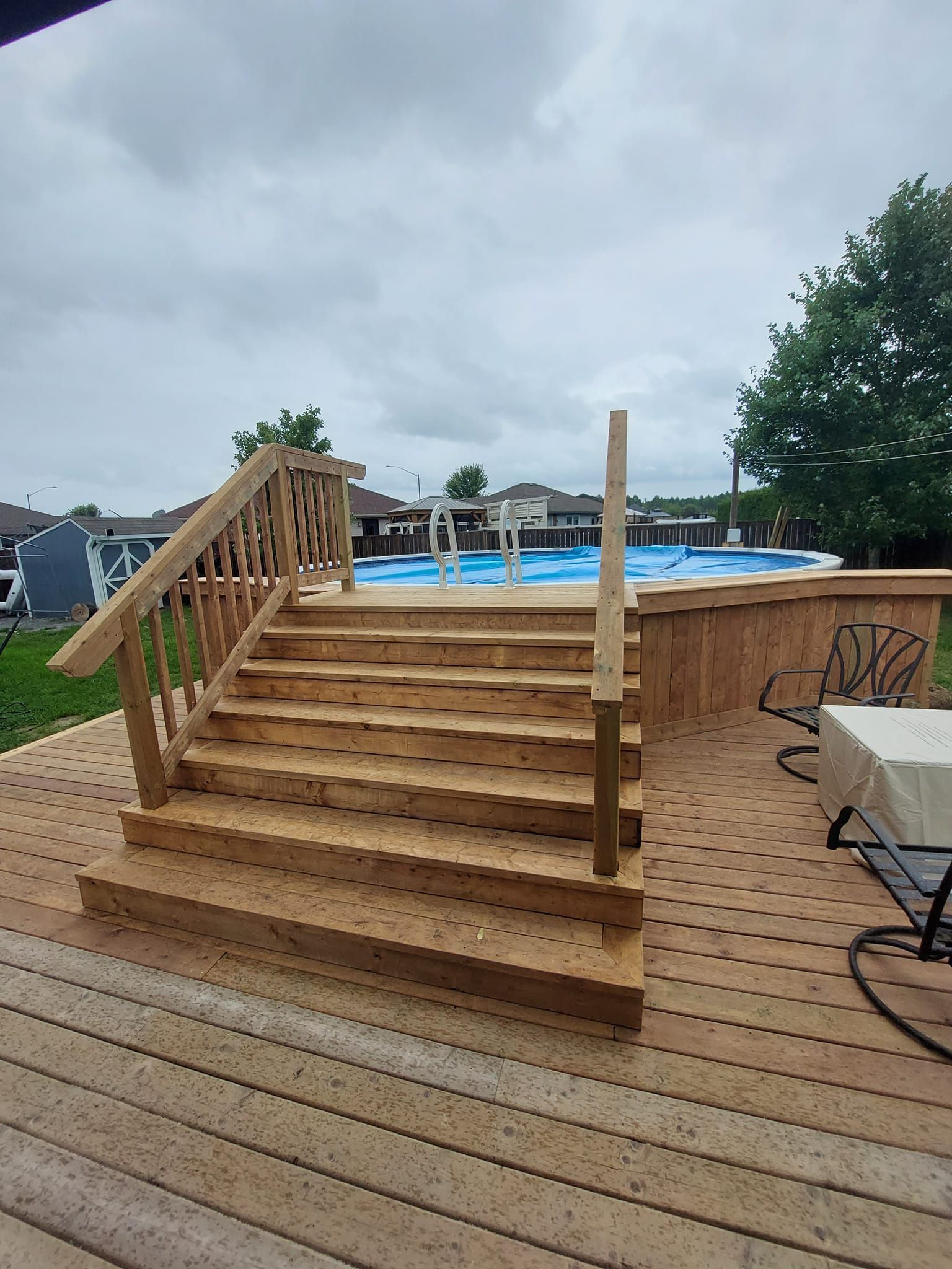 Wooden stairs leading up to an above-ground pool on a wooden deck. Cloudy sky overhead.