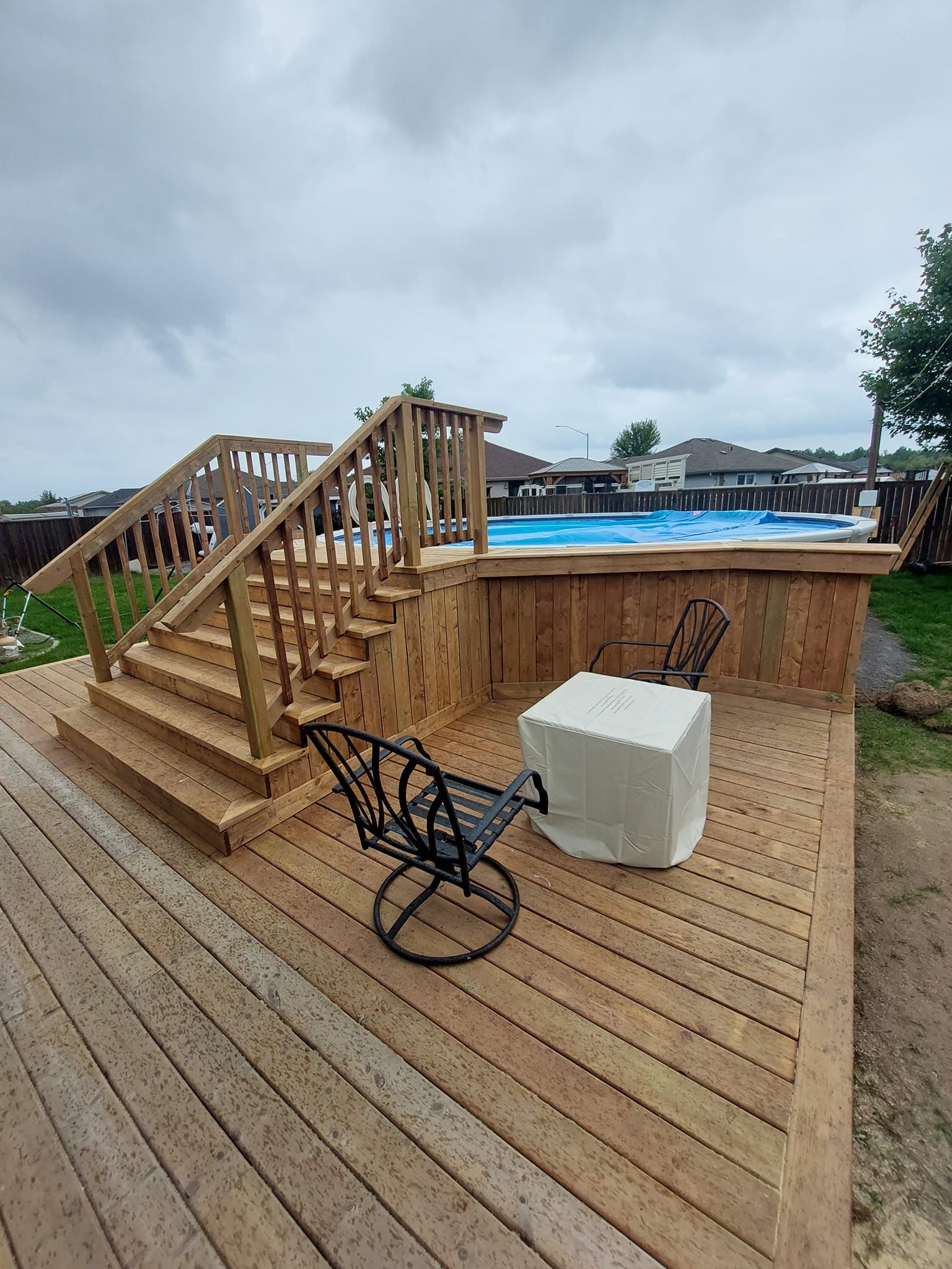 Wooden deck with stairs leading to a pool; two chairs and covered table. Cloudy sky.