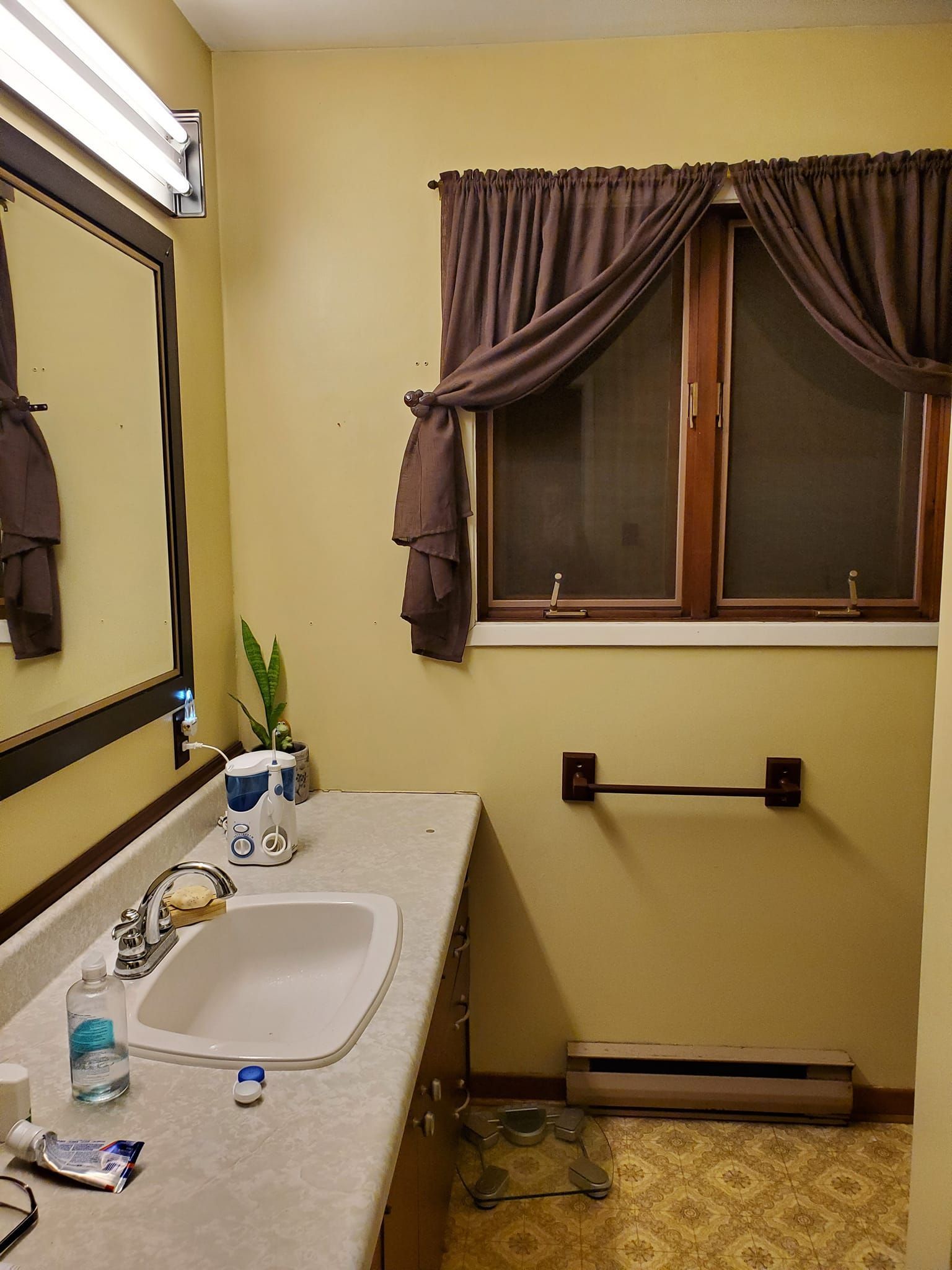 Bathroom with tan walls, brown window curtains, and a countertop with a sink and toiletries.
