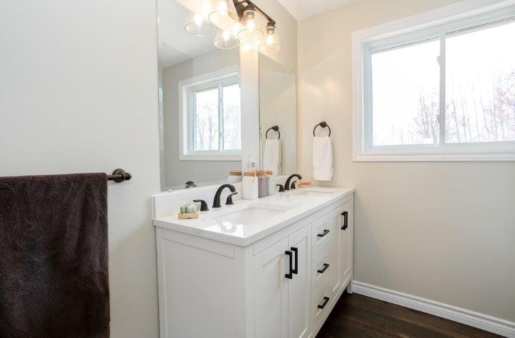 White bathroom with double vanity, mirror, and window; dark towel and trim.