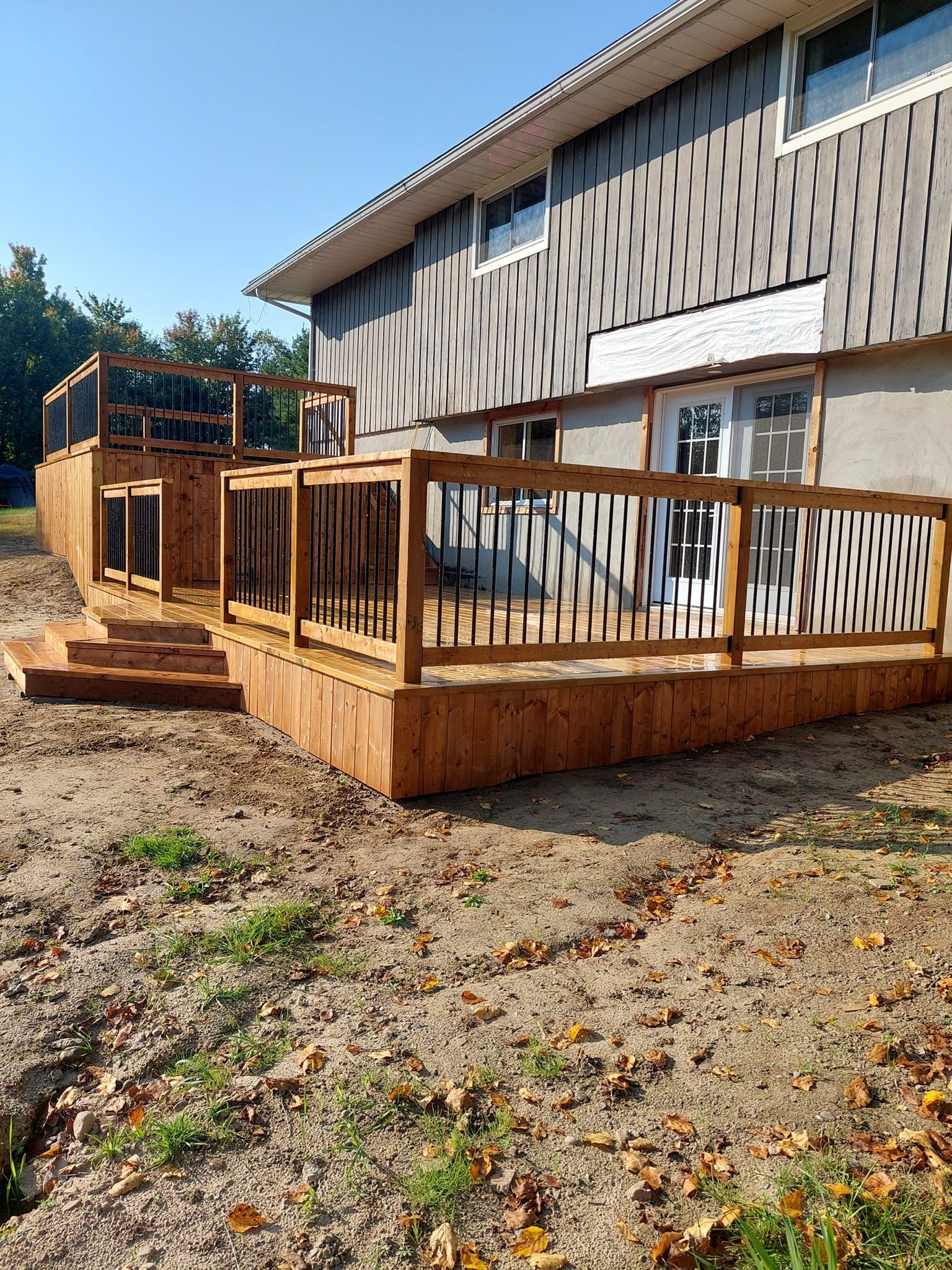 Wooden deck attached to a house with stairs and railings. Brown, black and tan colors with a blue sky.