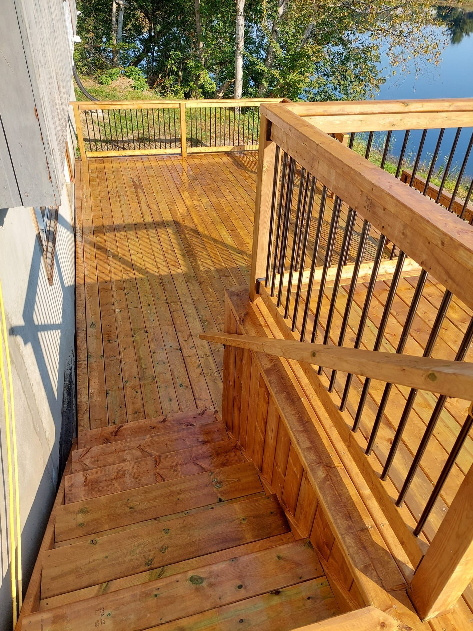 Wooden deck with stairs, railings, and black spindles, next to a building and overlooking water.