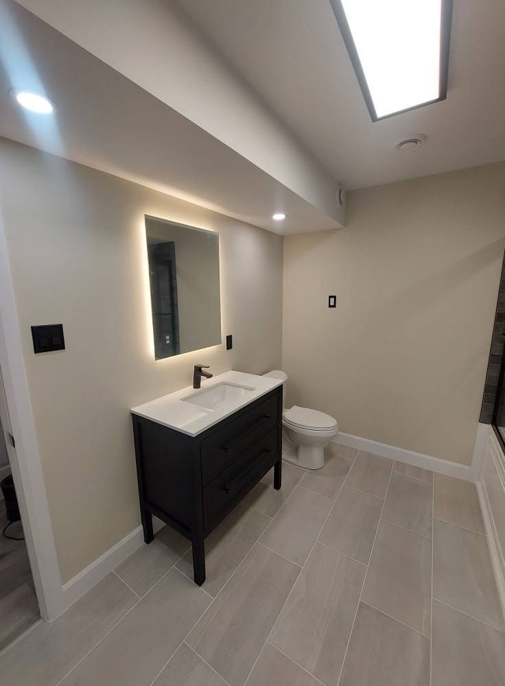 Bathroom with black vanity, lit mirror, white toilet, and gray tiled floor. Neutral walls, recessed lighting.