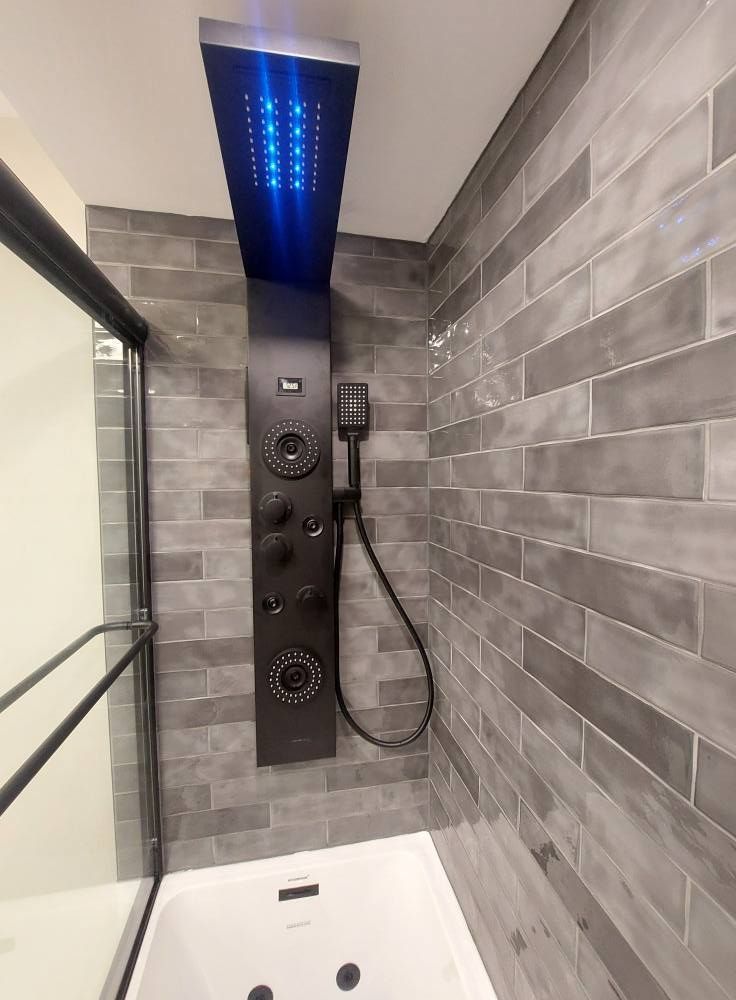 Black shower panel with controls and overhead LED lights against gray tiled wall.