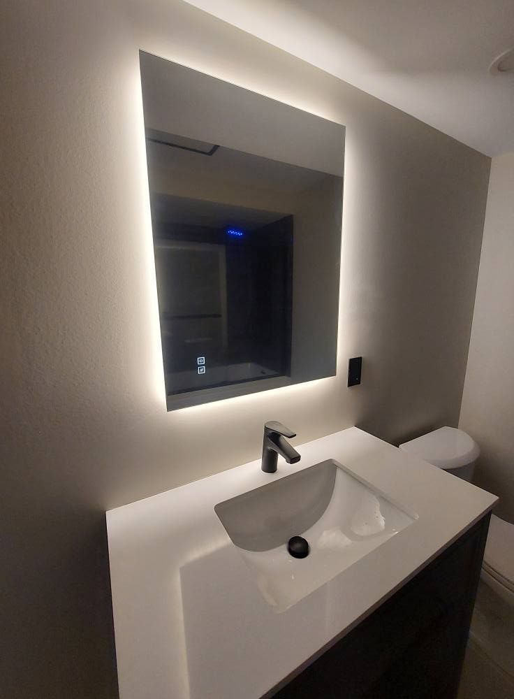 Modern bathroom with backlit mirror over a white countertop and sink, faucet, and toilet.