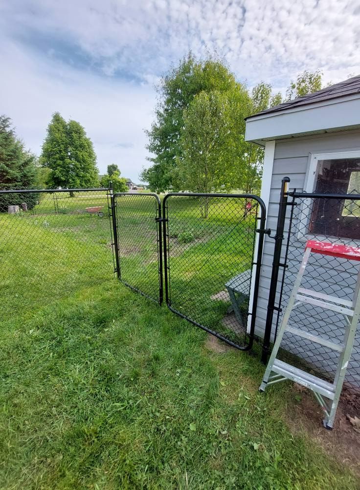 Black chain-link fence with gate, next to a shed. Green grass and trees under a blue sky.