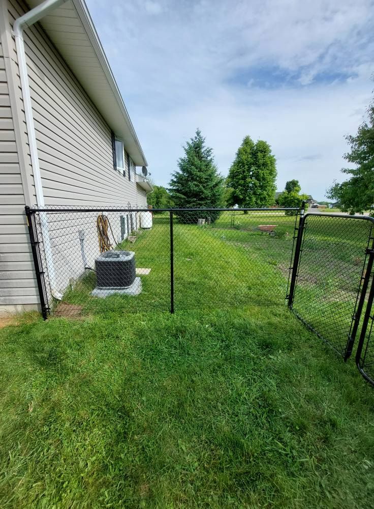 Black chain link fence surrounding a grassy backyard with an air conditioning unit near a light-colored house under a blue sky.
