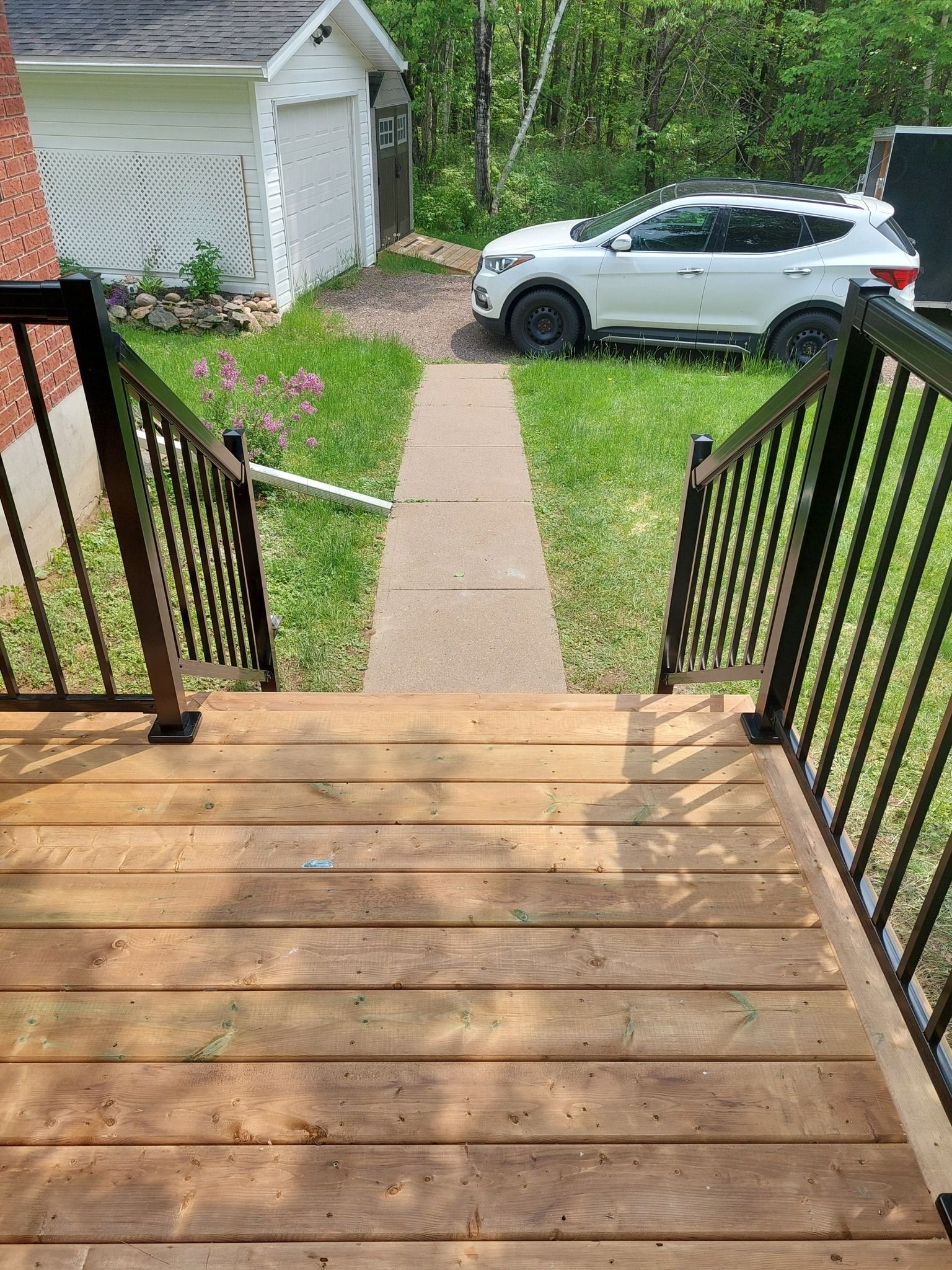 Wooden deck with black railing leads to a concrete path, lawn, car, and garage.