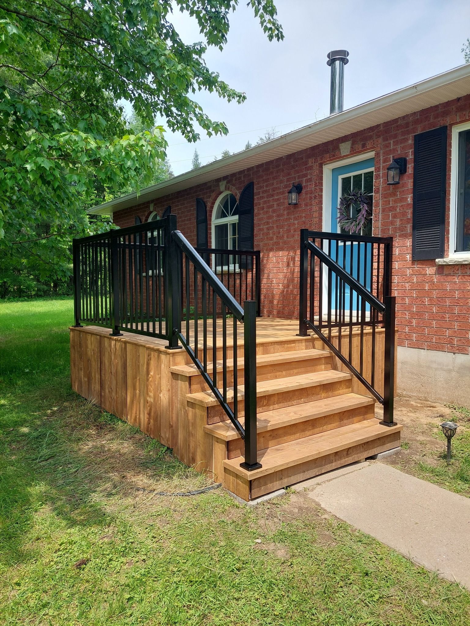 Wooden deck with black railing and stairs leading to a brick house with a blue door.