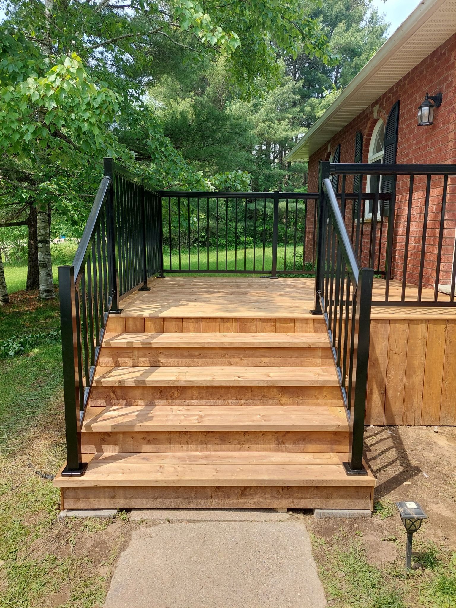 Wooden deck with black railing, steps up to a brick house, on a sunny day.