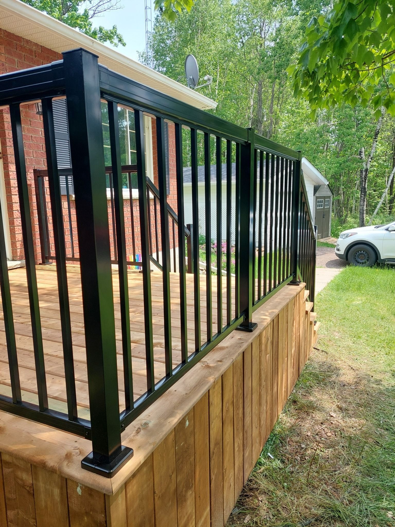 Black railing on a wooden deck outside a house with green trees and grass.