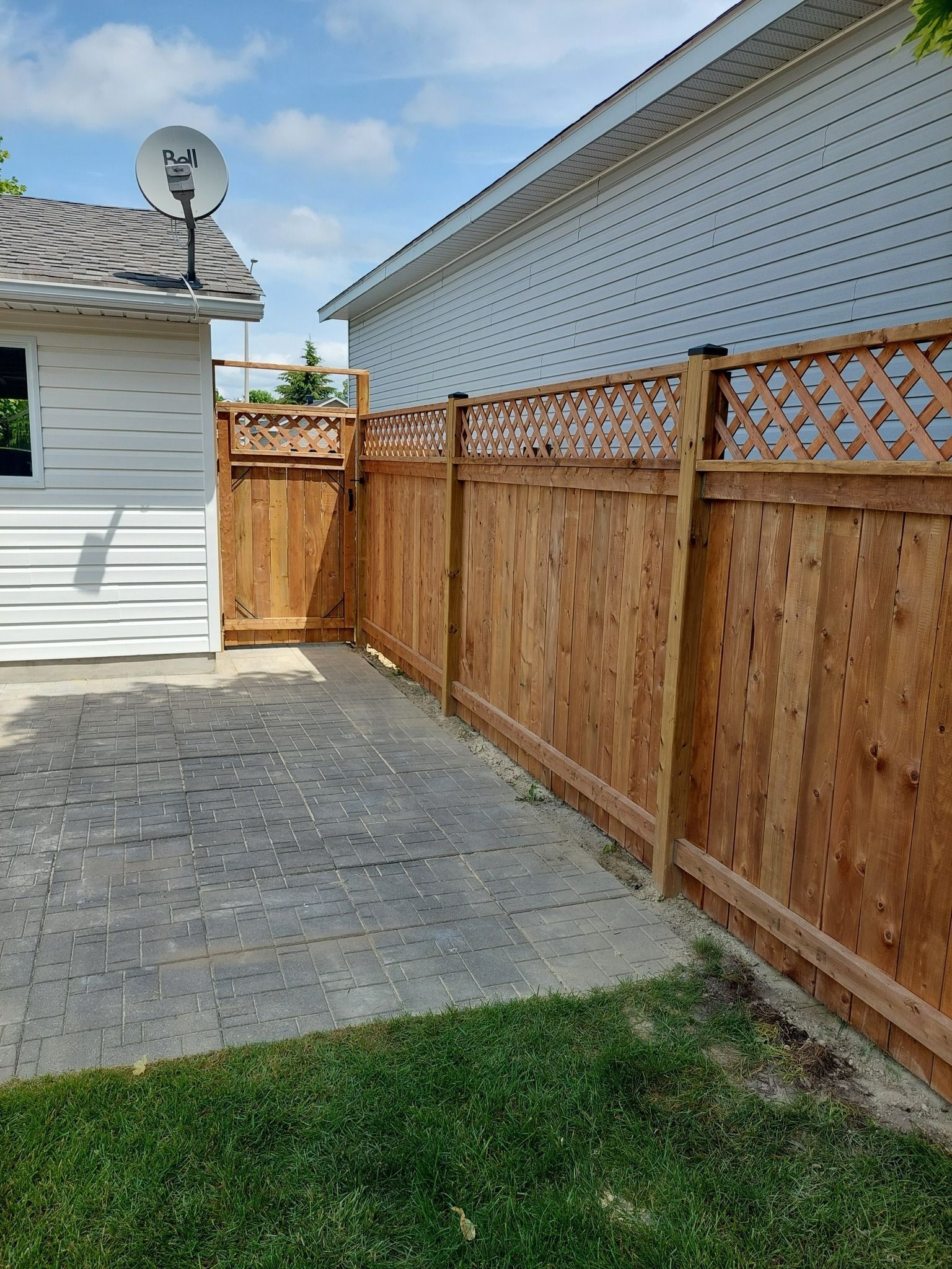 Brown wooden fence with lattice top borders a yard with a paved area and grass. A white building is on the left.