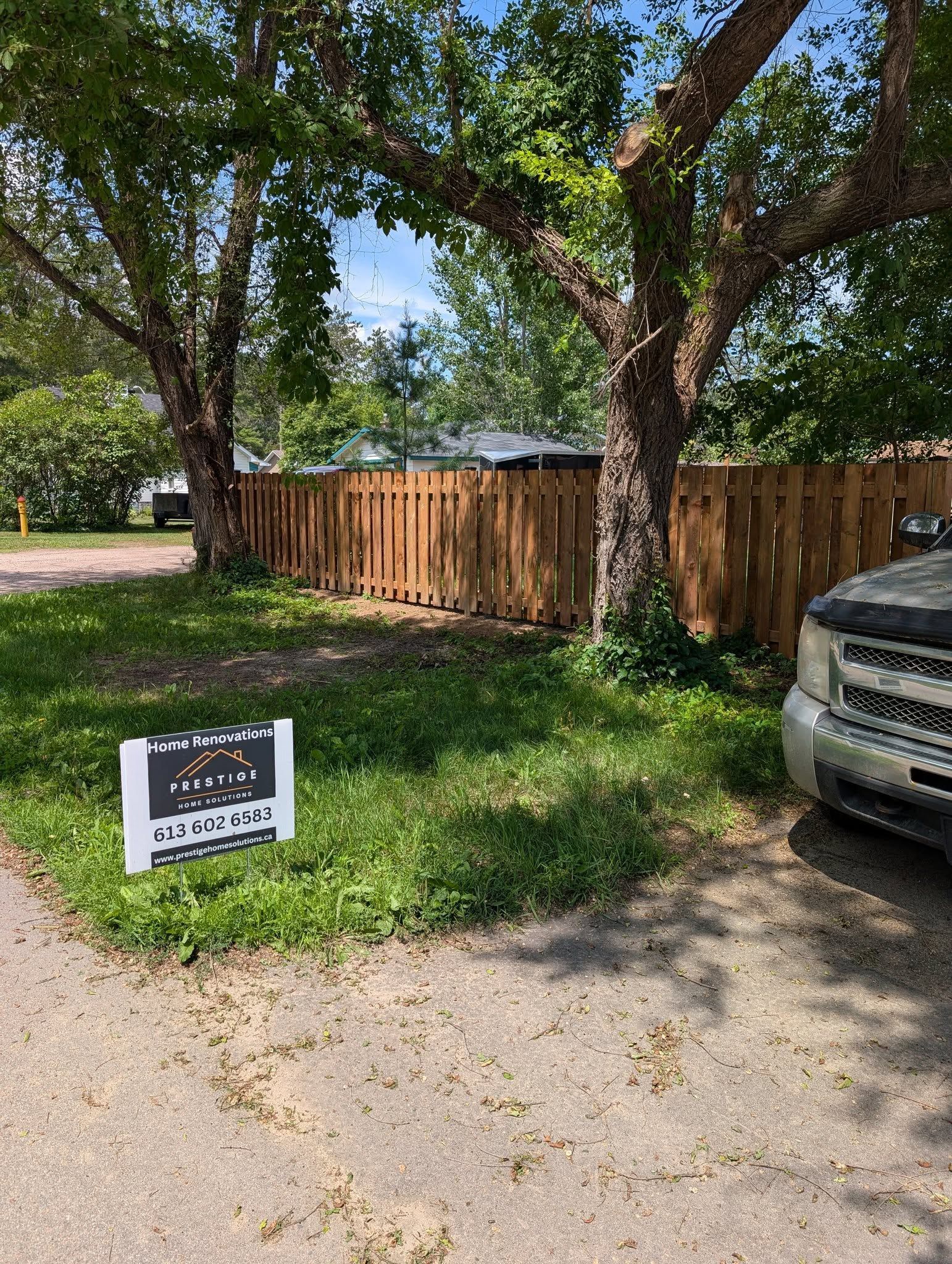 A wooden fence and two trees stand in front of a house on a sunny day. A sign sits in the grass.