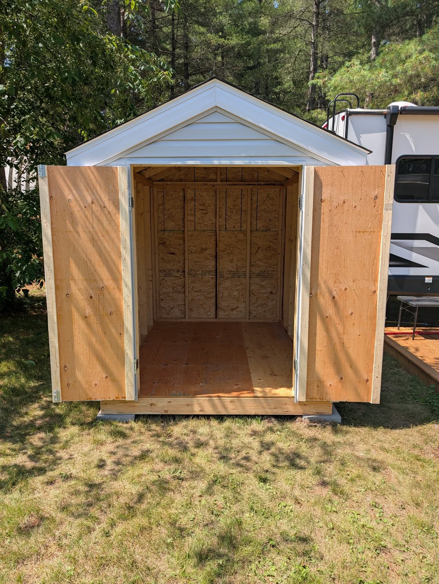 Small, unfinished wooden shed in a grassy yard, open doorway, light brown siding.