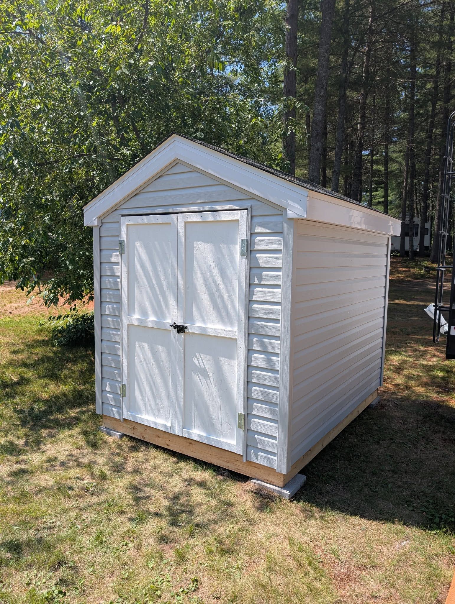 White shed with double doors in a grassy yard; trees in the background.