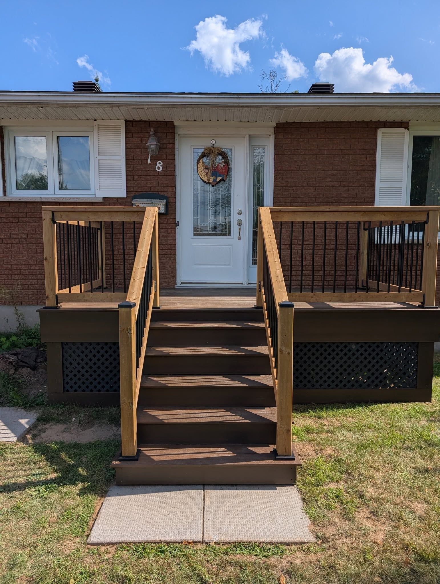 Brown wooden steps lead up to a white front door with a wreath. Brown house siding, deck, and lawn visible.
