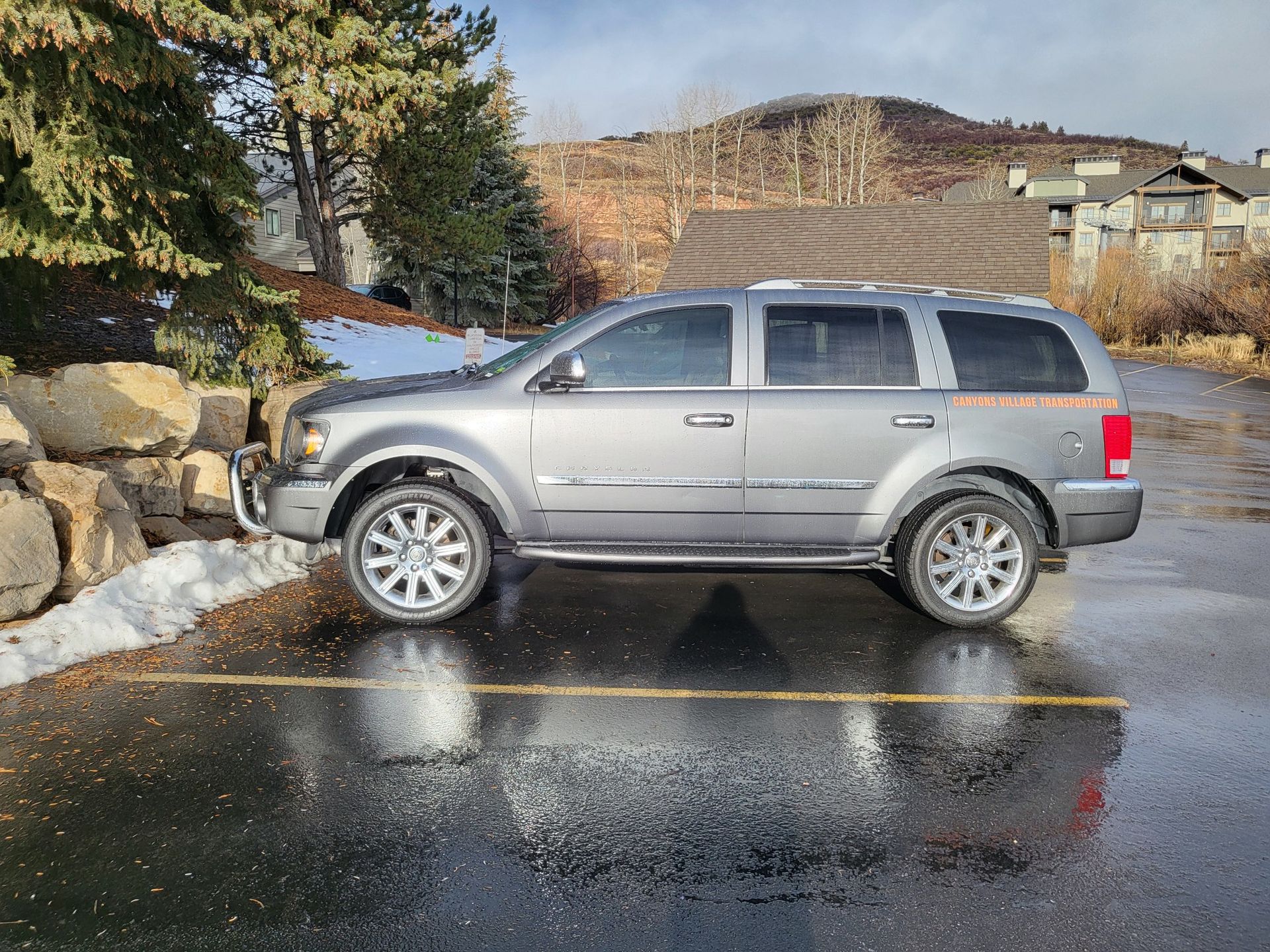 Silver SUV parked in a wet lot beside a stone wall and snowy trees
