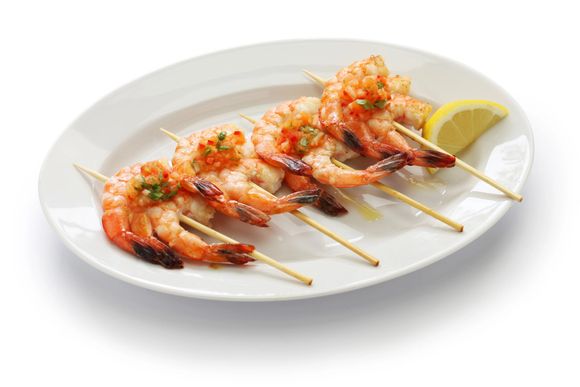 Un plato blanco cubierto con brochetas de camarones y una rodaja de limón.