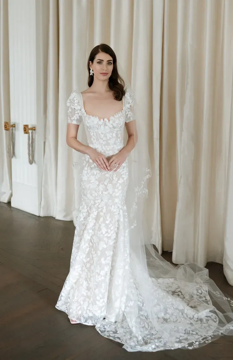 A woman in a white wedding dress is standing in front of a white curtain.
