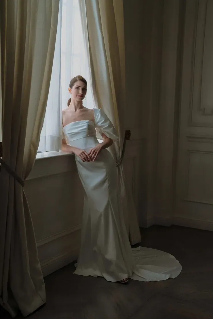 A woman in a wedding dress is leaning against a window.