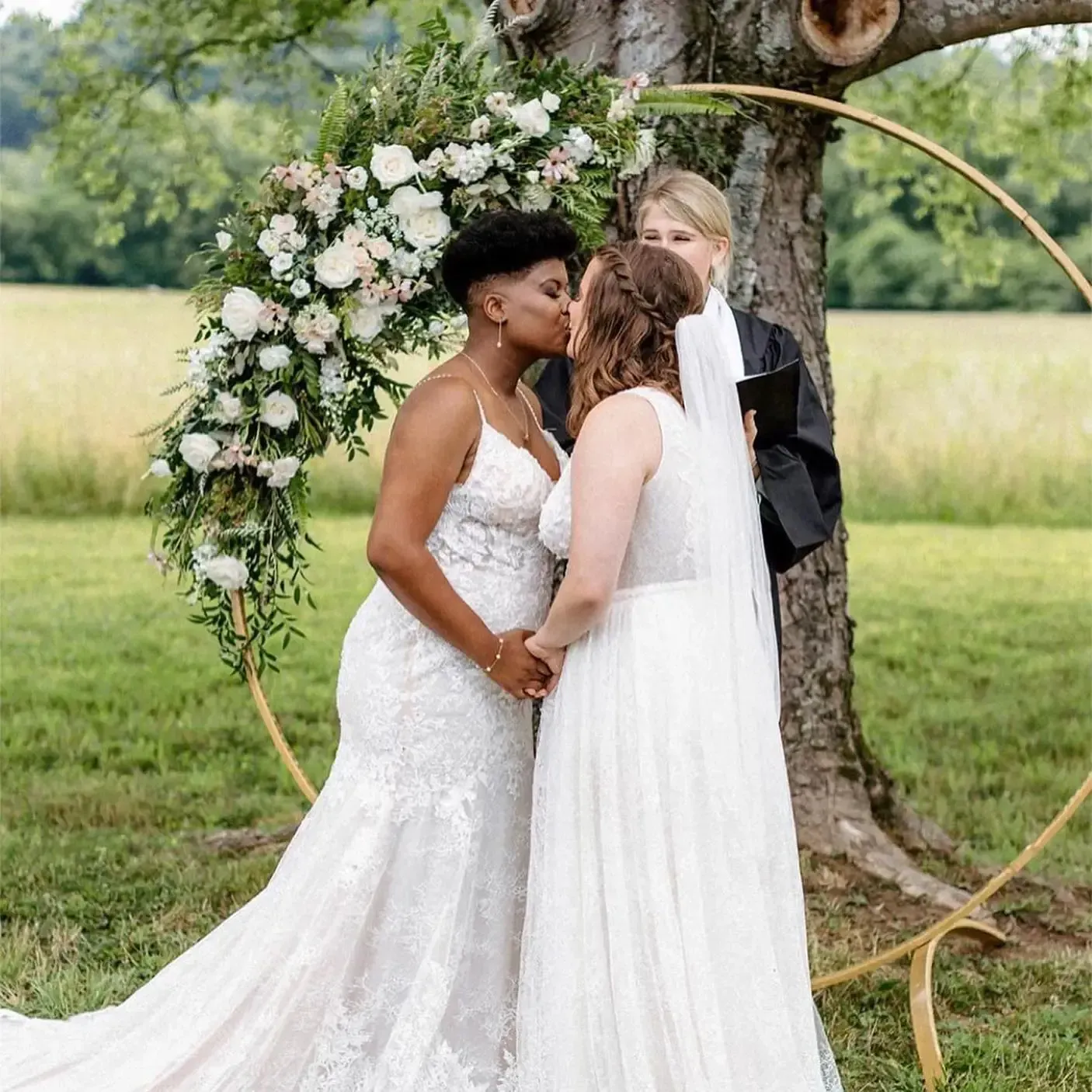 Two brides are kissing under a tree at their wedding ceremony.