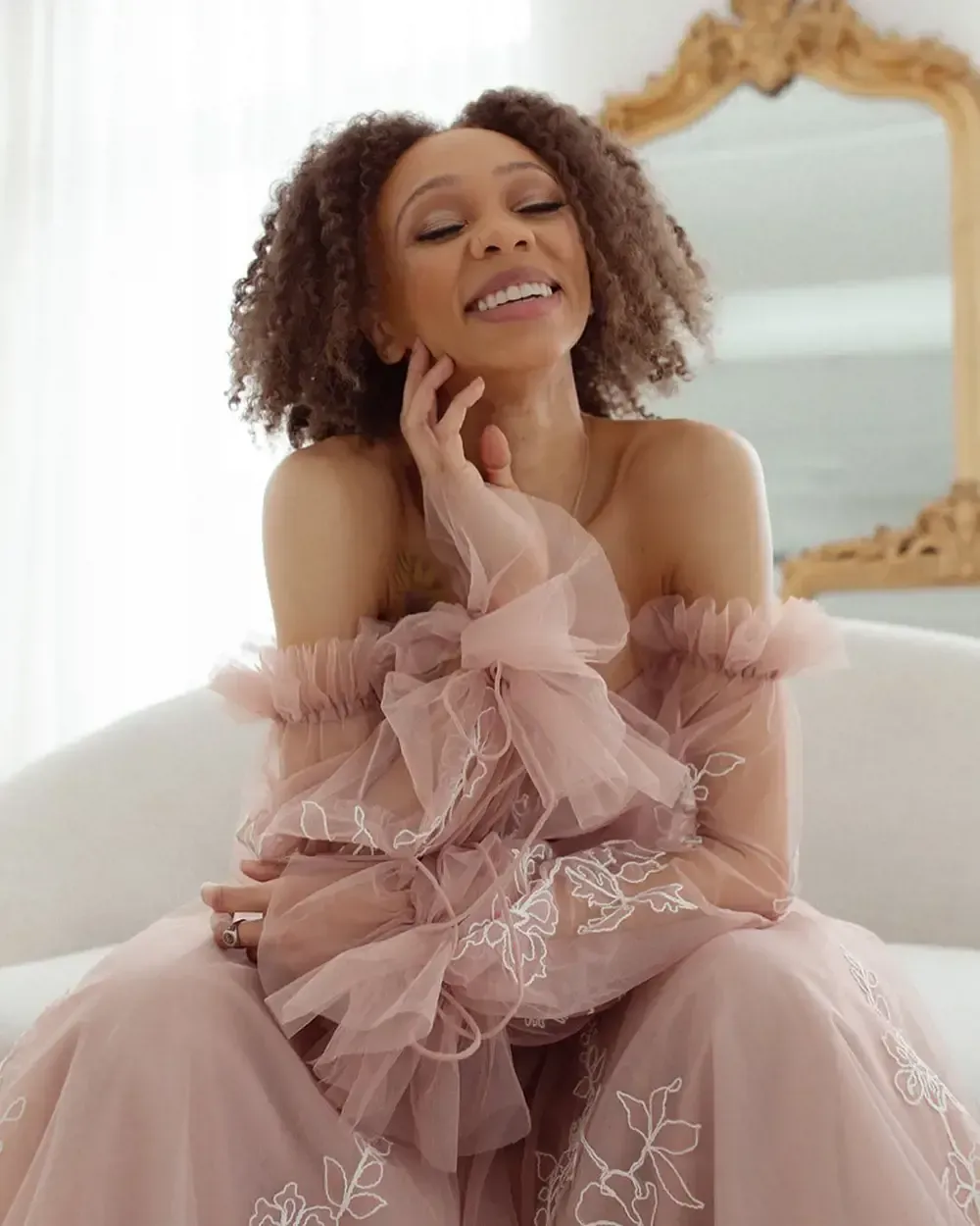 A woman in a pink dress is sitting on a couch in front of a mirror.