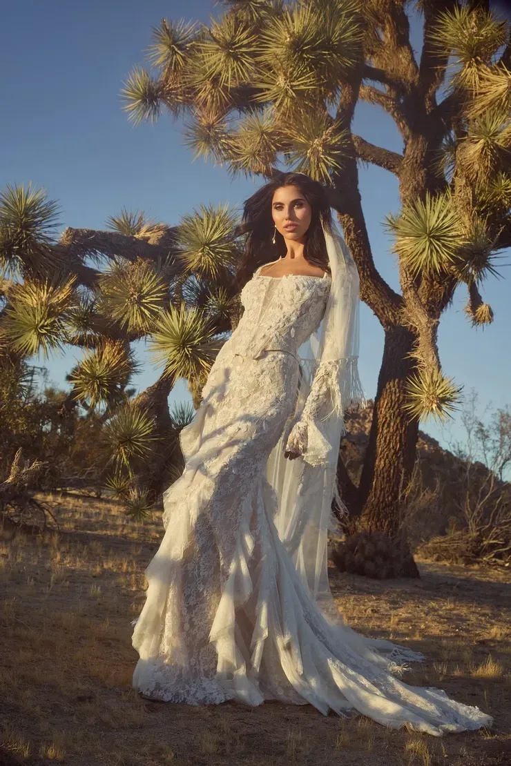 A woman in a wedding dress is standing in front of a tree in the desert.