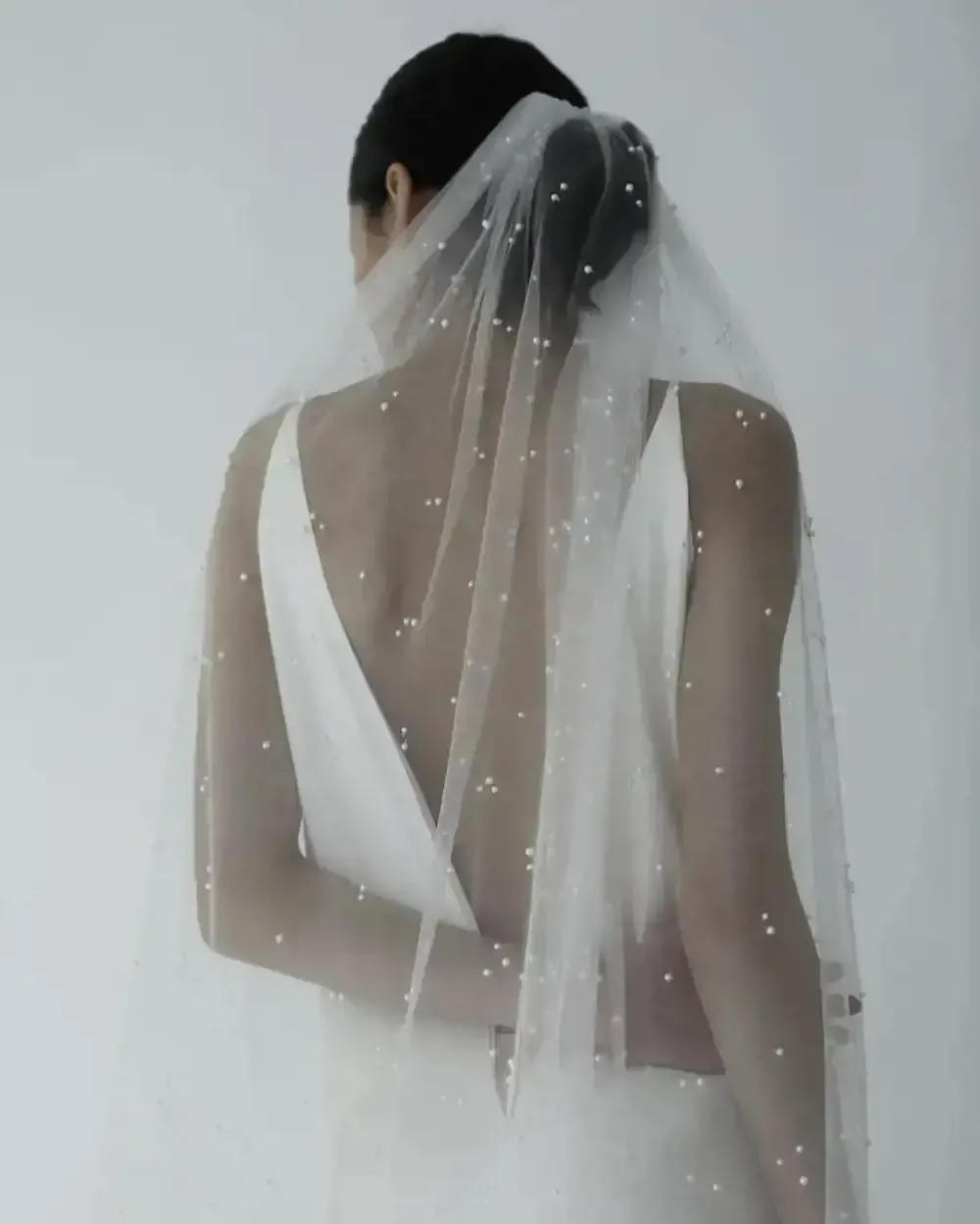 A woman in a white dress and veil is standing in front of a white wall.