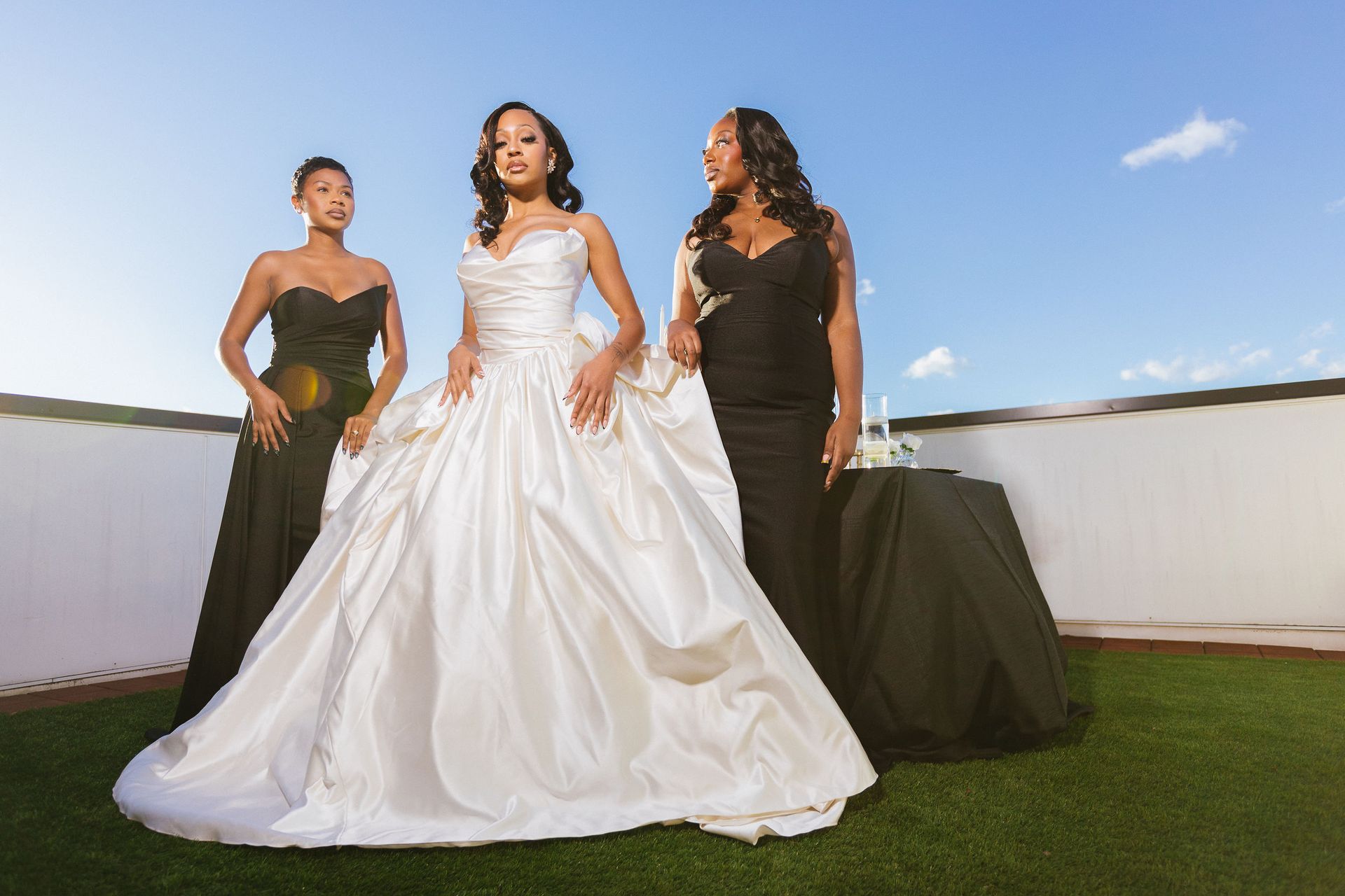 Bride in white gown with two bridesmaids in black dresses on a rooftop with clear sky.