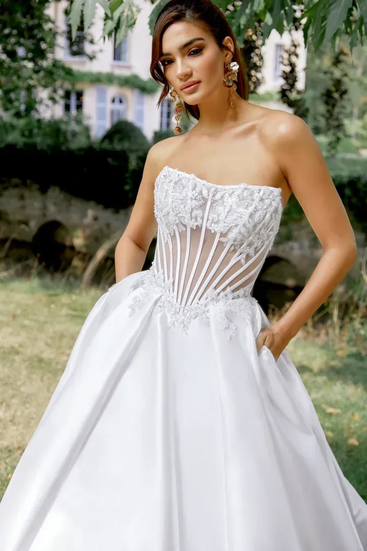 Woman in strapless white wedding dress with lace bodice, posing outdoors with hands in pockets.