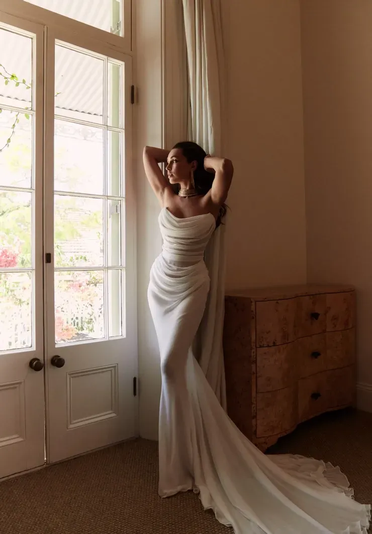 A woman in a wedding dress is standing in front of a window.