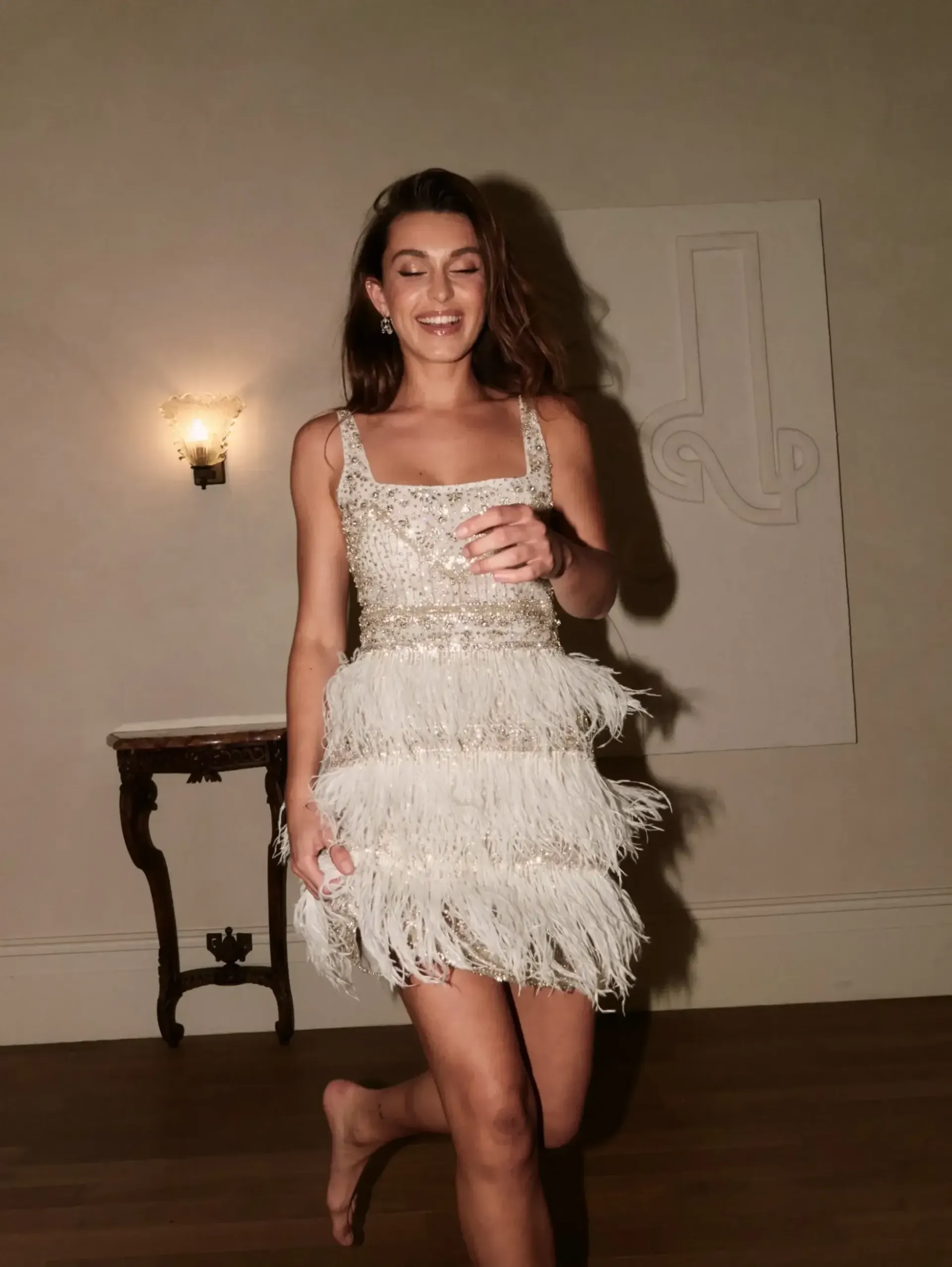 A woman in a white feathered dress is standing in a room holding a glass of wine.