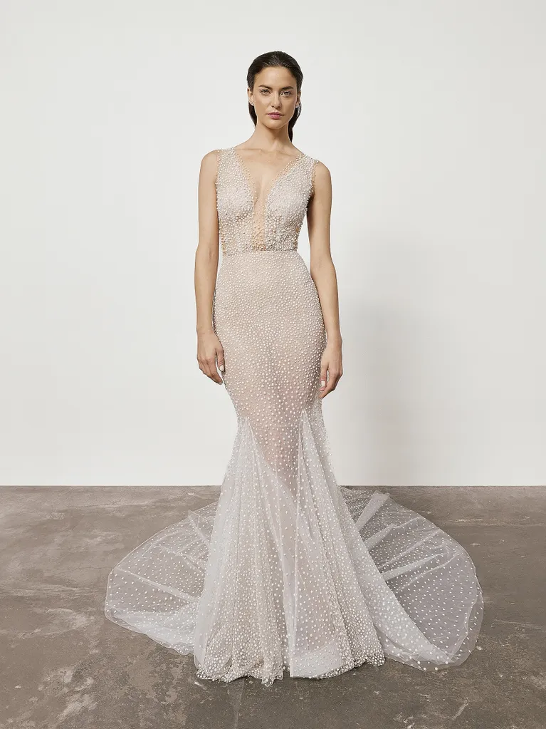 Woman in sheer, beaded, mermaid-style wedding gown with a long train, posing in a studio.