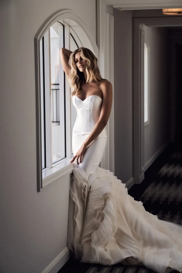 A woman in a wedding dress is standing in front of a window.
