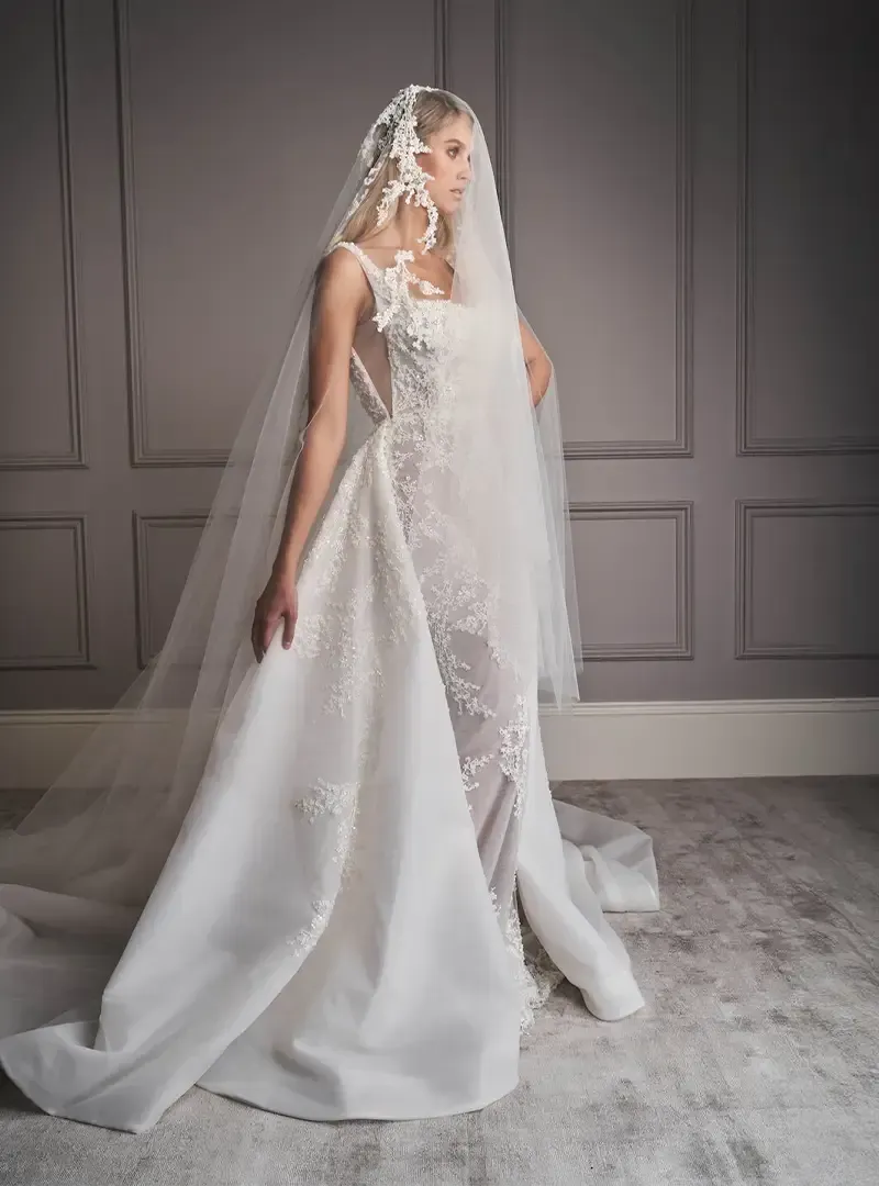 A woman in a white wedding dress and veil is standing in front of a wall.