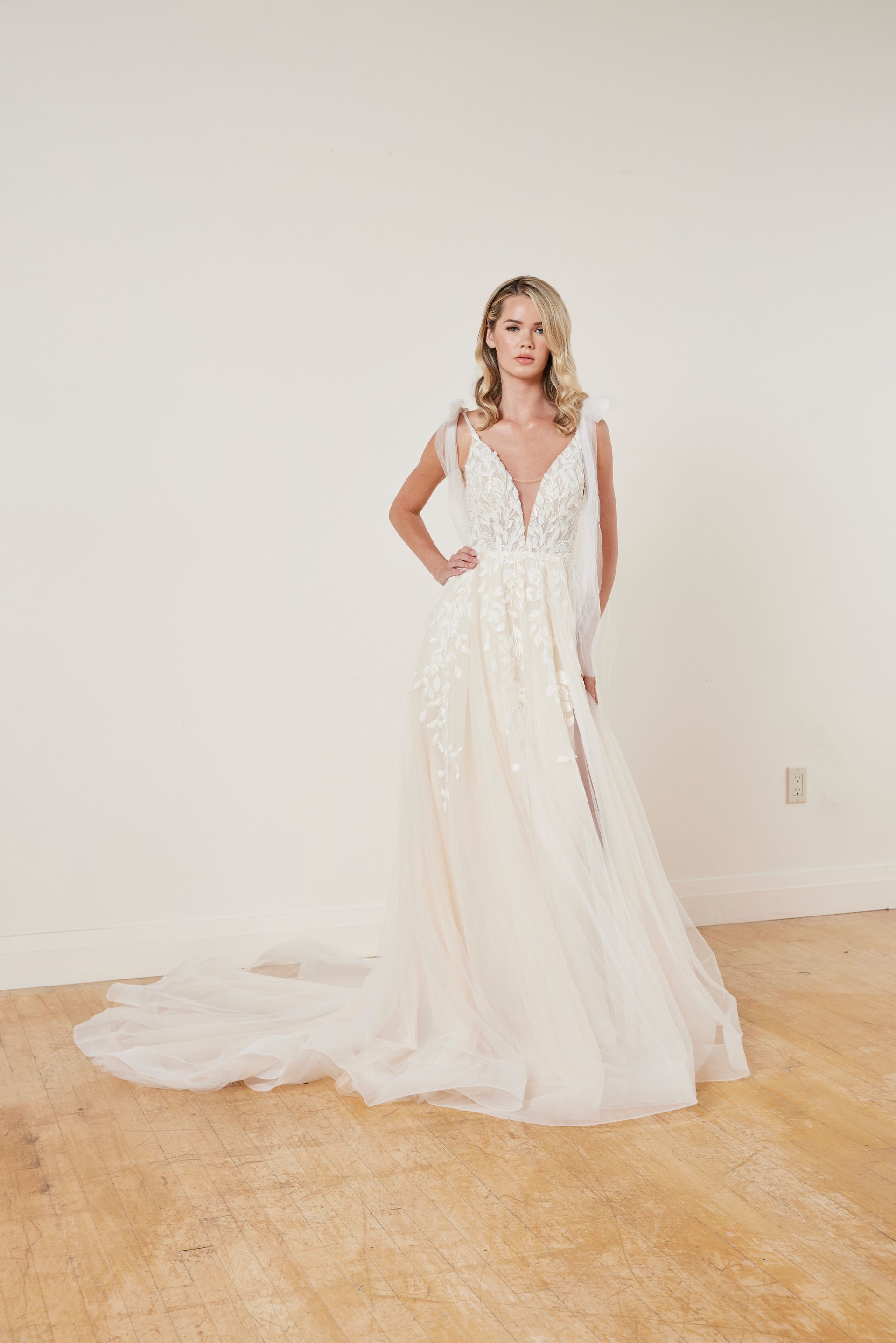 Woman in a short white wedding dress with floral embellishments and a long veil stands against a wall.