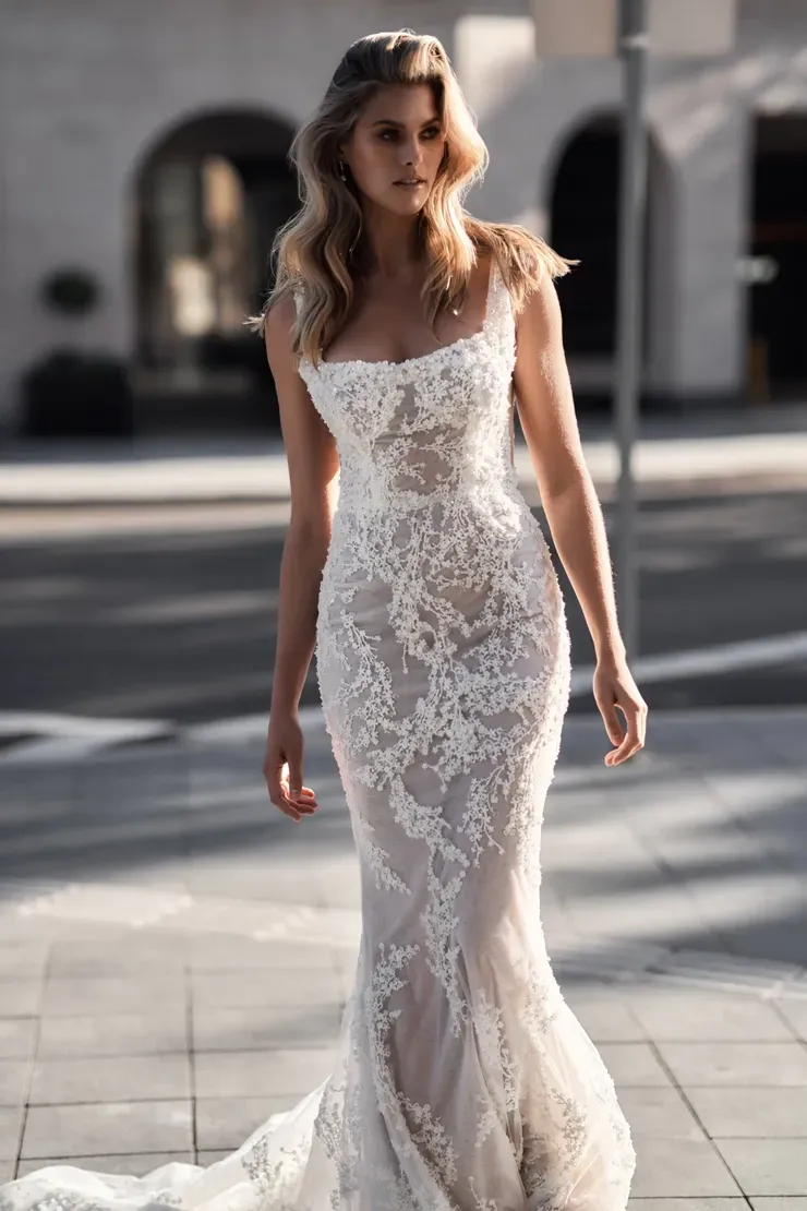 A woman in a white lace wedding dress is standing on a sidewalk.