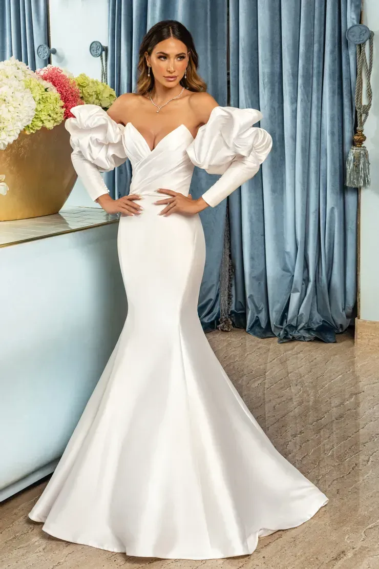 A woman in a white dress with ruffled sleeves is standing in front of a vase of flowers.