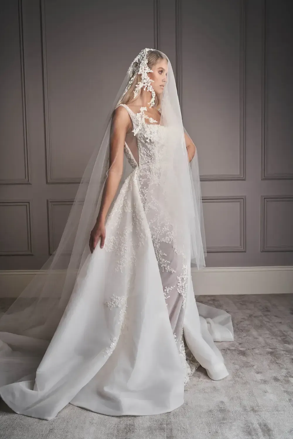 A woman in a white wedding dress and veil is standing in front of a wall.