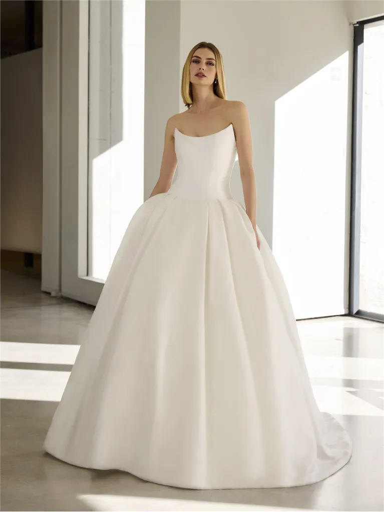 Woman in a white strapless ball gown, standing in a bright interior space with large windows.