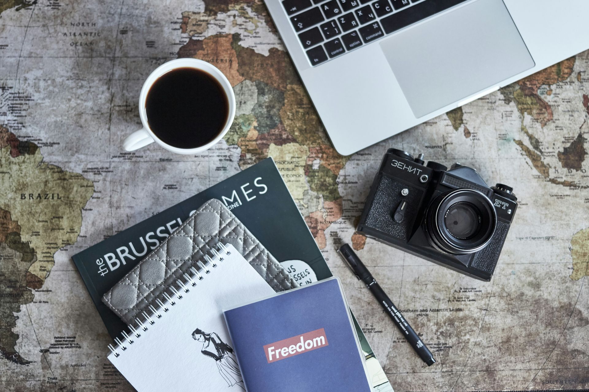 A cup of coffee , a notebook , a camera , and a laptop are on a table.