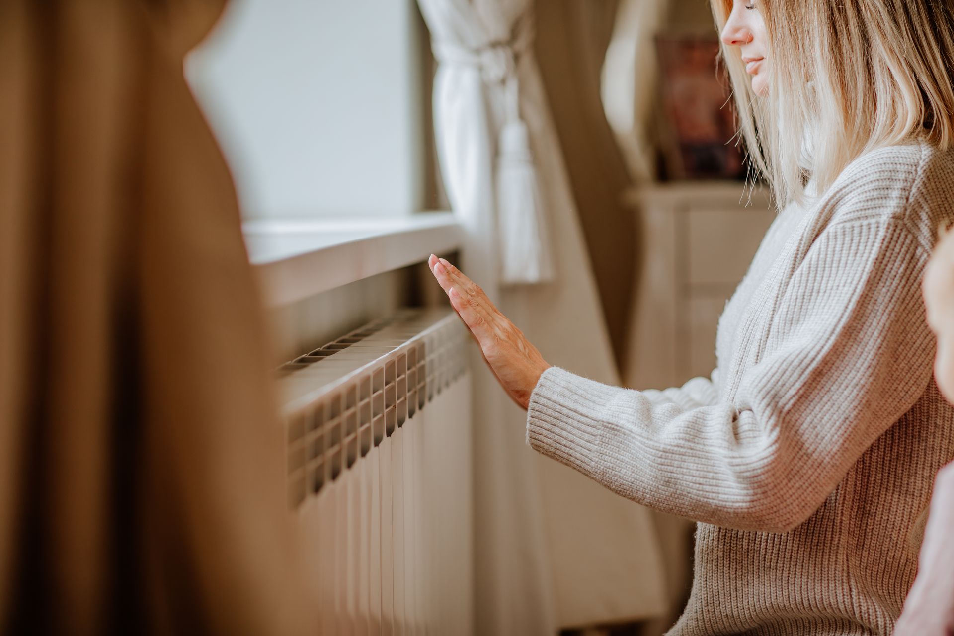 A woman in a long beige sweater is posing at home near the radiator.