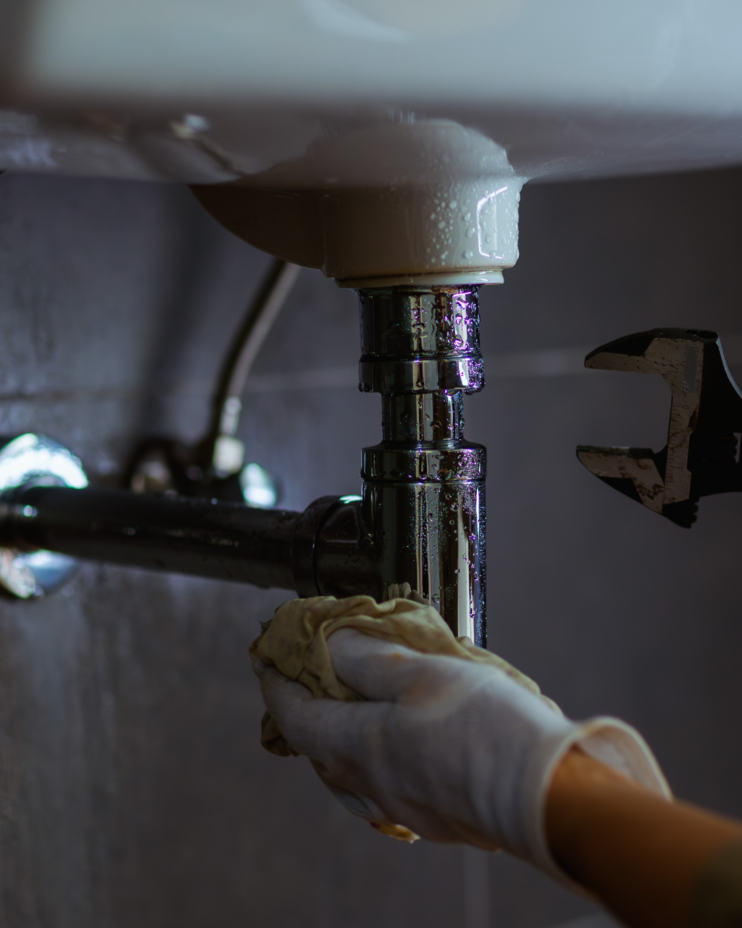 Une main gantée essuie l'eau d'un tuyau d'évier chromé tandis qu'une clé est tenue à proximité.