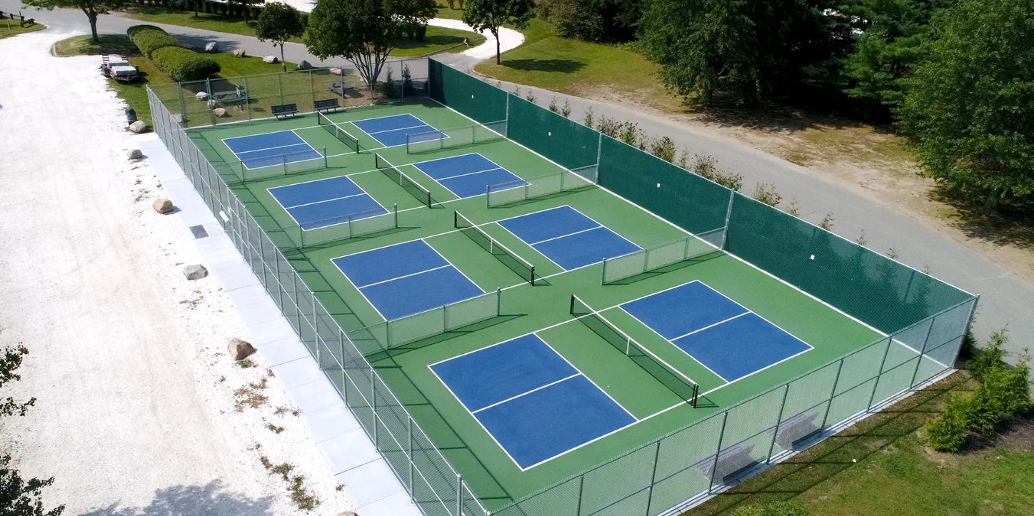 Aerial view of several pickleball courts, blue playing surfaces and green surrounding areas, enclosed by a fence.