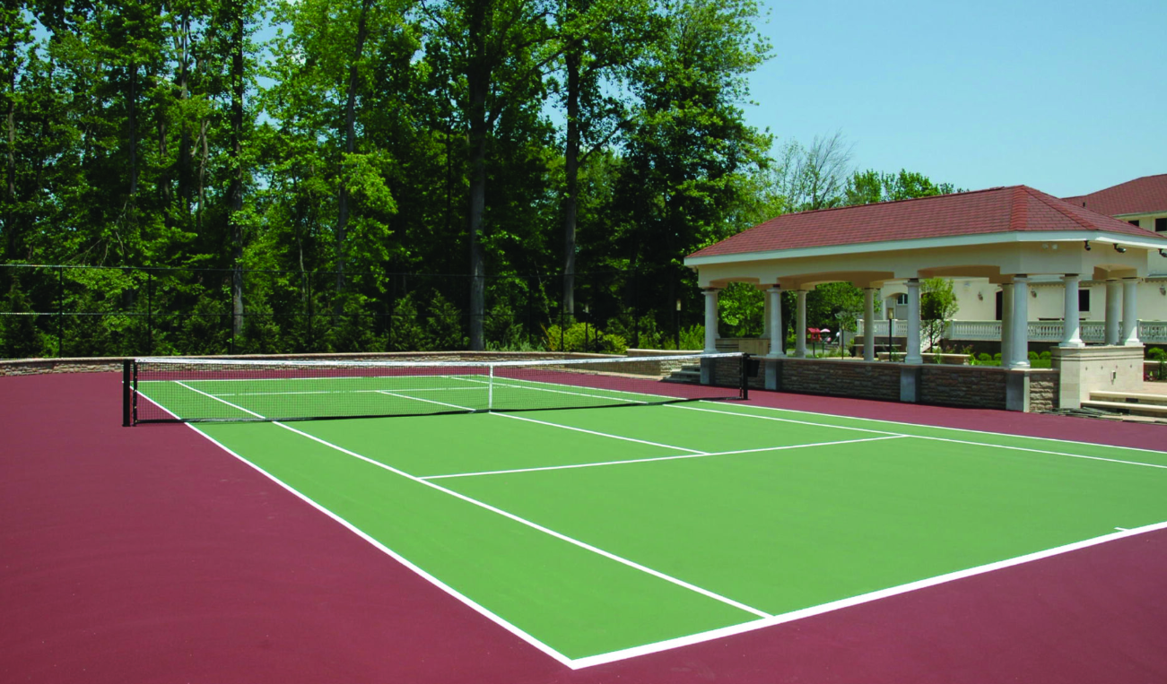 Tennis court with green playing surface and red surround, gazebo in background. Sunny day.