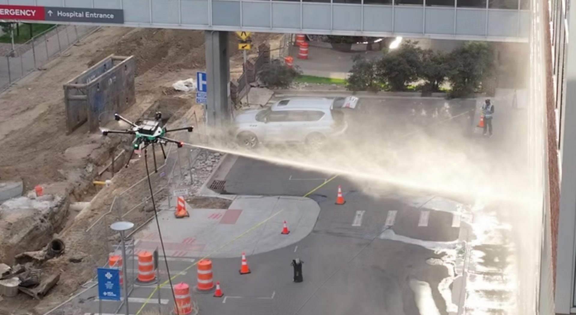 A drone sprays a mist onto a hospital building wall near a construction area with traffic cones and signage.