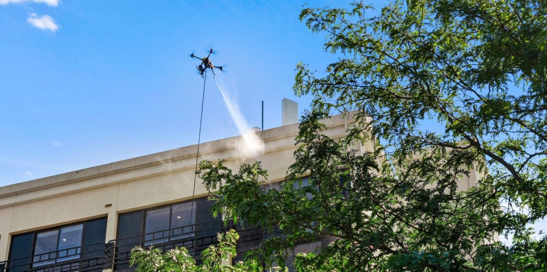 A drone tethered to the ground spraying a stream of water or cleaning solution onto the exterior wall of a building.
