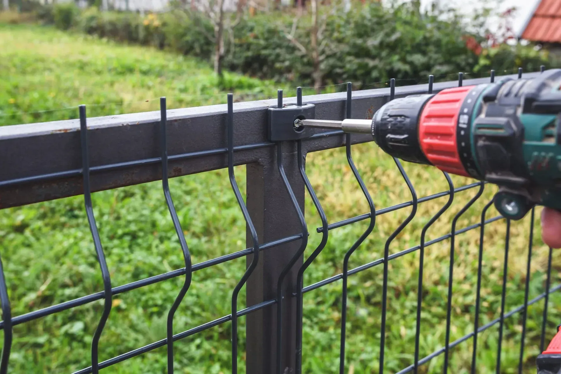 Person using a power drill to attach a black fence panel to a black post outdoors on a grassy lawn.